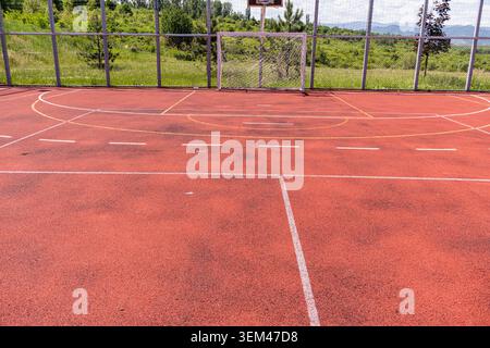 Ein horizontaler Außenrahmen eines Sportspielplatzes, der einen verwitterten roten Acrylboden mit geometrischen weißen Farbmarkierungen und einen Fußball zeigt Stockfoto