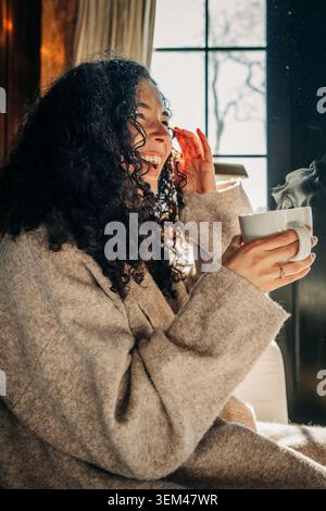 Lächelnde Frau genießt ein heißes Getränk an einem sonnendurchfluteten Fenster Stockfoto