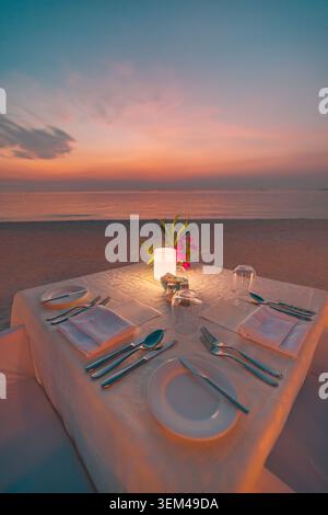 Romantischer Kerzenlicht-Esstisch neben ruhigem Meer bei pastellfarbenem Sonnenuntergang, elegante Tischdekoration. Tropischer Sommerabend, Hochzeitsreise. Luxusresort-Paar Stockfoto