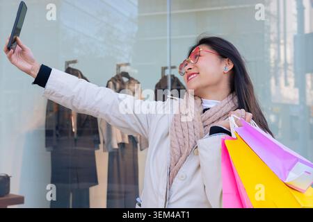 Lächelnde Frau, die Einkaufstaschen hält und draußen ein Selfie mit ihrem Smartphone macht. Stockfoto