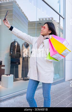 Lächelnde Frau, die Einkaufstaschen hält und draußen ein Selfie mit ihrem Smartphone macht. Stockfoto