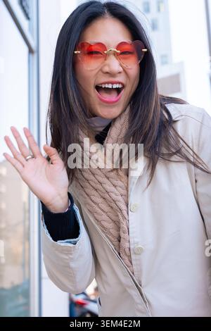 Die aufgeregte junge Frau winkte mit der Hand und lächelte, während sie eine herzförmige Brille trug. Stockfoto