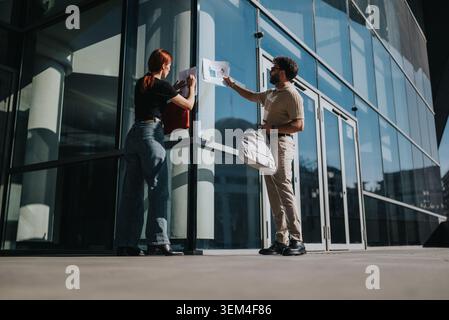 Arbeitsfreunde tauschen an einem sonnigen Tag Dokumente vor einem modernen Glasbürogebäude aus Stockfoto
