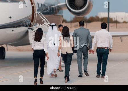 Multiethnische Geschäftsleute und traditioneller Partner aus dem Golf-Arabien gehen gemeinsam in Richtung Privatjet über den Flughafenparkplatz, von hinten gesehen Stockfoto