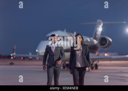 Zwei junge, selbstbewusste Geschäftsleute Mann im grauen Anzug und Frau im schwarzen Blazer gehen mit zielgerichtetem Schritt von weißen Privatjet-Flugzeugen Stockfoto
