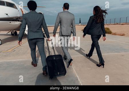 Selbstbewusstes multiethnisches Business-Team in Anzügen, das mit rollendem Gepäck in Richtung Privatjet über die sonnige Flughafenaspel geht, von hinten gesehen, ideal für Stockfoto