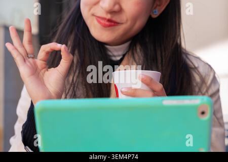 Lächelnde asiatische Frau, die OK Geste mit Tablet und Kaffee auf der Terrasse des Outdoor Cafe macht Stockfoto