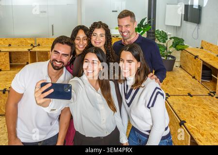 Eine Gruppe von sechs Kollegen posiert für ein Selfie in einem hellen, modernen Coworking Space. Sie lächeln und genießen ihre gemeinsame Zeit und zeigen Teamgeist. Stockfoto