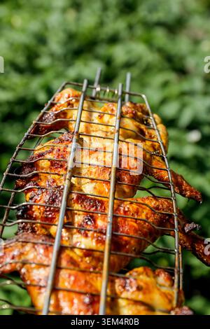 Nahaufnahme eines ganzen gegrillten Hühnchens in einem Metalldraht-Bratkorb draußen. Goldene knusprige Hähnchenflügel und Fleisch auf Grillrost mit verschwommenem Stockfoto