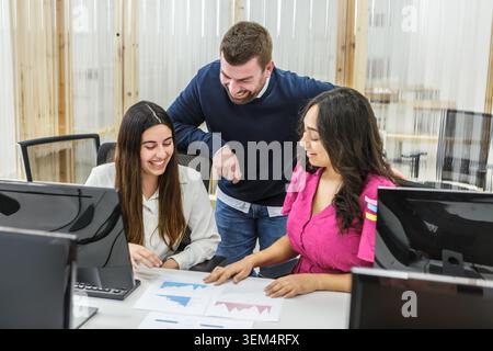 Drei Personen diskutieren, während sie gedruckte Diagramme in einem modernen Coworking-Raum überprüfen. Sie konzentrieren sich auf ihre Arbeit und arbeiten an ID zusammen Stockfoto