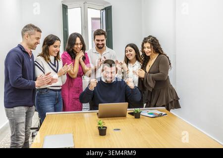 Eine Gruppe von sieben Personen ist in einem Coworking-Raum versammelt. Sie sehen sich eine Präsentation auf einem Laptop an. Ein Mann feiert mit Aufregung Stockfoto