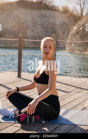 Eine Frau sitzt auf einer Matte am Wasserrand und übt an einem klaren, hellen Tag Sport. Stockfoto