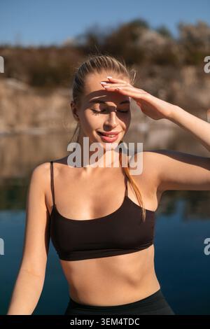 Eine junge Frau dehnt sich und trainiert am Wasser bei hellem Sonnenlicht, während sie ihre Augen mit der Hand abschirmt. Stockfoto
