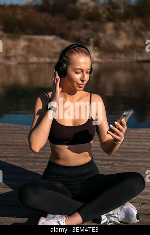 Eine Frau sitzt im Kreuz an einem See, während sie Musik auf ihrem Telefon hört. Es ist tagsüber und sonnig. Stockfoto