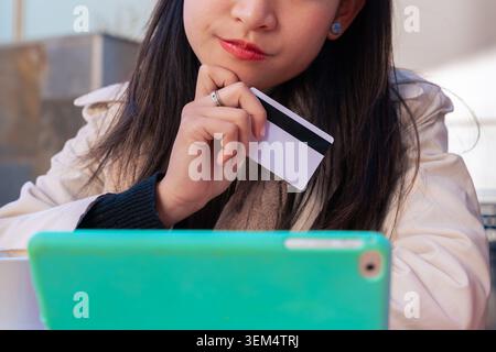 Nachdenkliche asiatische Frau mit Kreditkarte und Tablet am Schreibtisch im Freien im Café Stockfoto