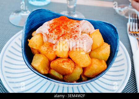 Eine Schüssel mit spanischen Patatas Bravas, knusprigen gebratenen Kartoffelwürfeln, serviert mit scharfer Sauce und Paprika, steht auf einem Restauranttisch zum Teilen bereit Stockfoto
