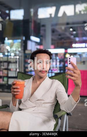 Ein vertikales Premium-Porträt eines Influencers, der einen nicht-binären Schwulen aus Thailand zeigt, der einen Orangeneistee oder ein boba-Getränk hält, während er ein Smartphone benutzt Stockfoto