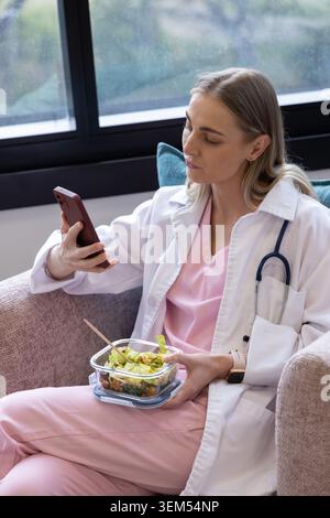 Weibliche Ärztin in rosafarbenen Peelings, die Stethoskop trägt und das Telefon hält, während sie Salat auf dem Sofa isst Stockfoto