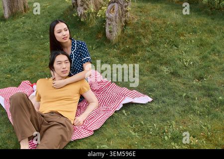Liebevolles asiatisches Paar sitzt zusammen auf einer Picknickdecke, umgeben von der Natur unter sonnigem Himmel. Stockfoto