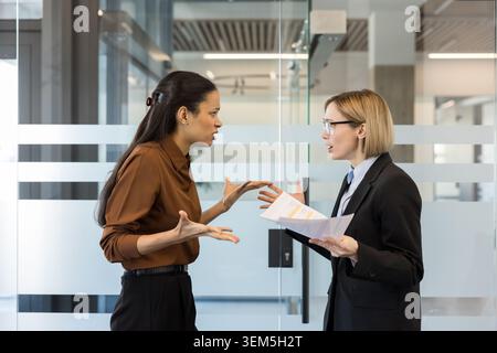 Zwei Geschäftsfrauen streiten während eines angespannten Unternehmensmeetings über Papierkram und zeigen Frustration, Meinungsverschiedenheiten und Stress, während Kollegen Zeugen eines hitzigen Streits am Arbeitsplatz sind Stockfoto