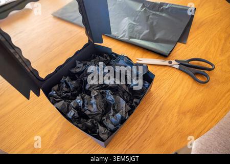 Zerknittertes Tissuepapier in der offenen matten schwarzen Box in der Nähe einer Schere auf einem Tisch. Konzept: Handgefertigte Geschenkverpackung Stockfoto