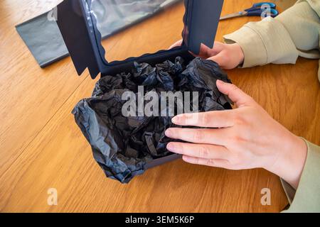 Hände legen zerknittertes Taschentuch in eine offene, matte schwarze Box auf einem Tisch. Konzept: Handgefertigte Geschenkverpackung Stockfoto