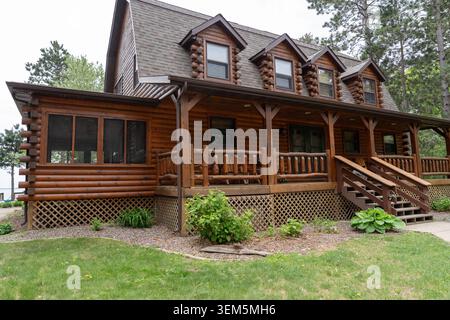 Ein wunderschönes horizontales Foto eines luxuriösen zweistöckigen Holzblockhauses mit einer überdachten Veranda, rustikalen Holzgeländern, Holztreppen und Dormer wi Stockfoto