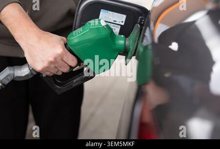 Ein detailliertes Nahfoto, das die Hände eines Mannes einfängt, einer trägt ein Ehering, hält eine grüne Kraftstoffpumpendüse und steckt sie in das offene Fu Stockfoto