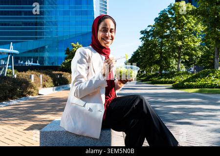 arabische Geschäftsfrau aus dem Nahen Osten im Hijab, die durch die Stadt schlendert - lächelnde Frau mit Hijab, die einen frischen Salat vor einem modernen Bürogebäude genießt Stockfoto