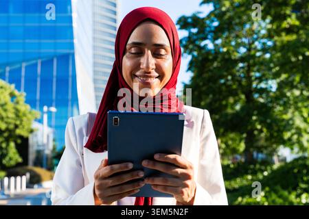 arabische Geschäftsfrau aus dem Nahen Osten im Hijab, die durch die Stadt schlendert – muslimische Geschäftsfrau lächelt, während sie im Freien ein digitales Tablet verwendet Stockfoto