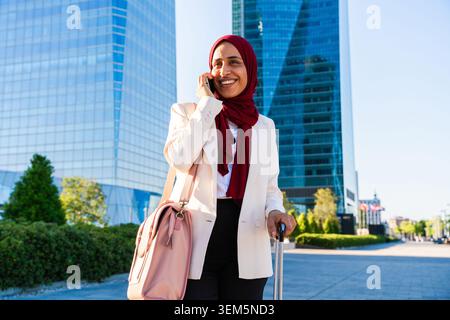 Eine arabische Geschäftsfrau aus dem Nahen Osten, die in der Stadt einen Hijab trägt, spaziert durch die Stadt - eine glückliche muslimische Frau, die mit dem Gepäck unterwegs ist und am Telefon in einem städtischen Gebäude spricht Stockfoto