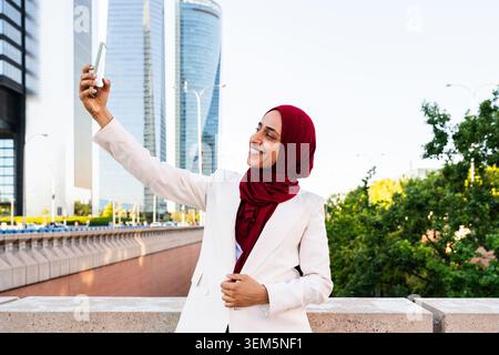 arabische Geschäftsfrau aus dem Nahen Osten im Hijab, die durch die Stadt schlendert - muslimische Geschäftsfrau lächelt, während sie ein Selfie mit einem Smartphone im Modus macht Stockfoto