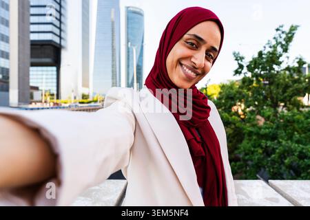 arabische Geschäftsfrau aus dem Nahen Osten im Hijab, die durch die Stadt schlendert - lächelnde Frau in Hijab und weißem Blazer, die ein Selfie mit einem Wolkenkratzer macht Stockfoto