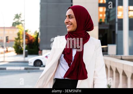 arabische Geschäftsfrau aus dem Nahen Osten im Hijab, die durch die Stadt schlendert - lächelnde junge arabische Frau in Hijab und weißem Blazer in einer Stadtkulisse Stockfoto