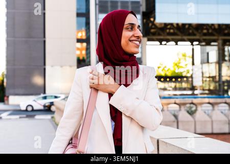 arabische Geschäftsfrau aus dem Nahen Osten im Hijab, die durch die Stadt schlendert - junge muslimische Geschäftsfrau im Hijab, die in einer urbanen Umgebung wegschaut Stockfoto