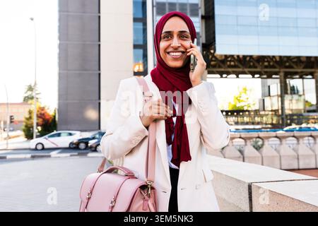 arabische Geschäftsfrau aus dem Nahen Osten im Hijab, die durch die Stadt schlendert - lächelnde Frau im Hijab, die am Telefon spricht, im urbanen Setting eine Aktentasche trägt Stockfoto