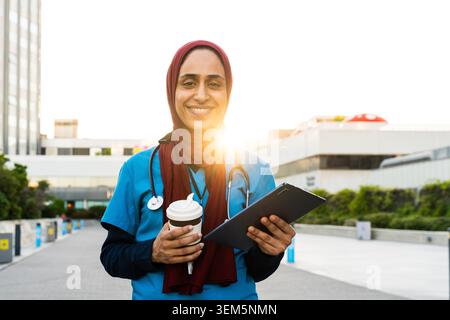Selbstbewusste arabische Ärztin, die einen Hijab und ein Stethoskop trägt, lächelt, während sie ein Klemmbrett und eine Kaffeetasse zum Mitnehmen vor einem modernen Krankenhausbu hält Stockfoto