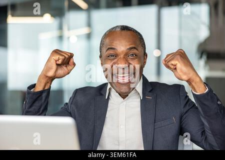 Ein reifer Unternehmensleiter jubelt mit den Fäusten an seinem Laptop, feiert einen Geschäftsgewinn und fühlt sich selbstbewusst, stolz und begeistert in einem modernen Büroumfeld Stockfoto