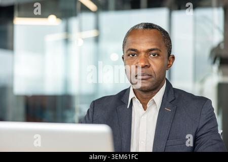 Reifer Mann mit ernsthaftem Ausdruck, der in die Kamera schaut, während er am Schreibtisch in einem Büro sitzt und Professionalität und Führung in einem Unternehmensumfeld darstellt Stockfoto