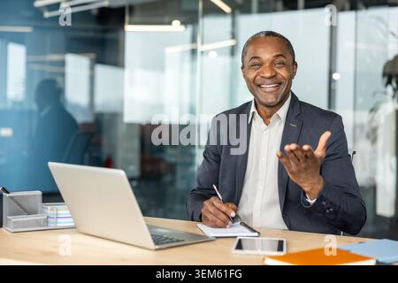 Reifer Geschäftsmann lacht und gestikuliert, während er an einem Schreibtisch mit Laptop und Notebook in einem modernen Büro sitzt und erfolgreiche Kommunikation und Führung repräsentiert Stockfoto