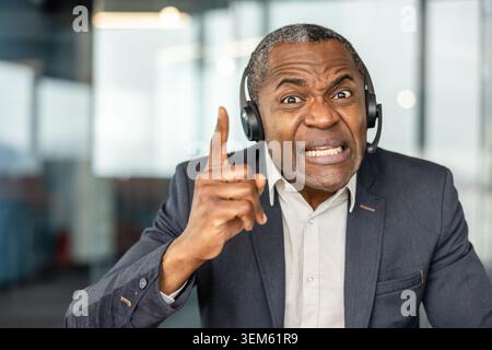 Reife Person, die in einem Callcenter arbeitet, ein Headset trägt und einen frustrierten Ausdruck zeigt, während sie mit dem Finger zeigt, was Wut, Stress oder ein schwieriges Gespräch darstellt Stockfoto