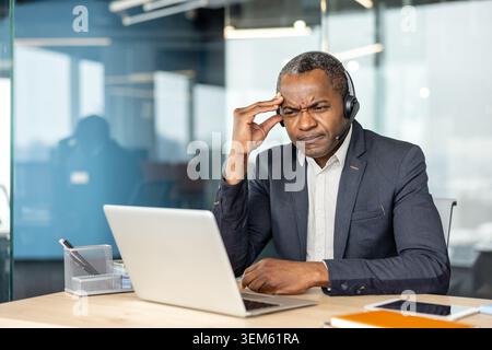 afroamerikanischer männlicher Vertriebsmitarbeiter mit Headset und Anzugsjacke, der an einem Schreibtisch sitzt und Stress und Frustration durch Kopfschmerzen erlebt, während er an einem Laptop arbeitet Stockfoto
