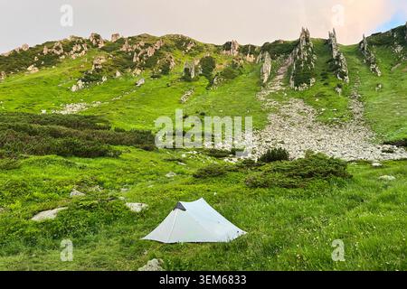 Einsames weißes Touristenzelt im üppigen grünen Tal, umgeben von majestätischen Hügeln und felsigen Klippen. Der Himmel oben zeigt einen Hauch von Sonnenaufgangsfarben und schafft eine ruhige Campingszene. Karpaten, Ukraine. Stockfoto