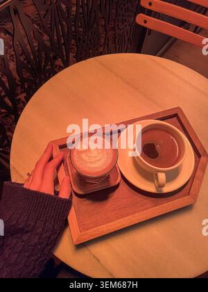 Blick auf die Hand einer Person, die auf einem Marmortisch neben zwei Tassen Kaffee auf einem Holztablett unter warmer oranger Beleuchtung in Beylikduzu, Istanbul, Turk, liegt Stockfoto
