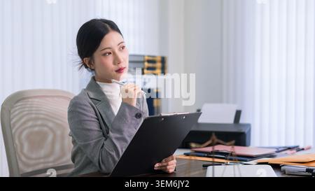Junge asiatische Anwältin, die eine Zwischenablage hält und Rechtsdokumente überprüft, arbeitet professionell und modern in der Büroumgebung Stockfoto