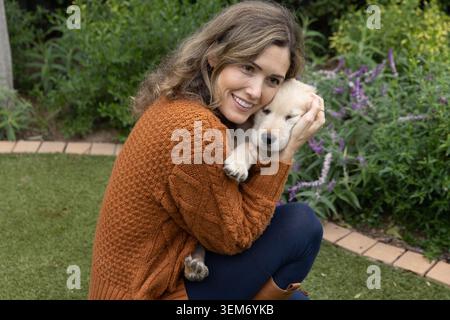 Mittelerwachsene Frau, die sich auf dem Rasen im Garten hockt, einen hellen Welpen umarmt, einen Rostpullover trägt Stockfoto