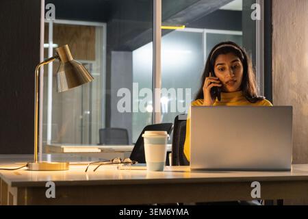 Indische Frau mit gelbem Rollkragen, die ein Smartphone hält und im Büro an silbernem Laptop arbeitet Stockfoto