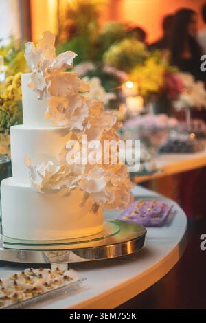 Blick auf eine dreistöckige weiße Hochzeitstorte mit zarten Zuckerblumen auf einem Dessertteller mit Blumenarrangements Rio de Janeiro, Bundesstaat Rio Stockfoto