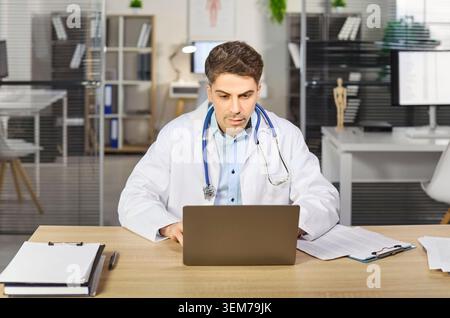Fokussierter Arzt mit Stethoskop am Laptop im modernen Klinikinnenraum Stockfoto