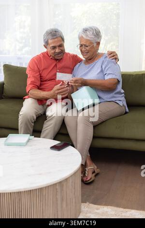 Seniorenpaar sitzt auf grünem Sofa im Wohnzimmer und öffnet hellblaue Geschenk- und Lesekarte Stockfoto